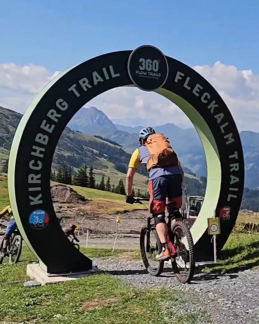 Biker fährt durch das Starttor des Kirchberg Trails mit Bergpanorama