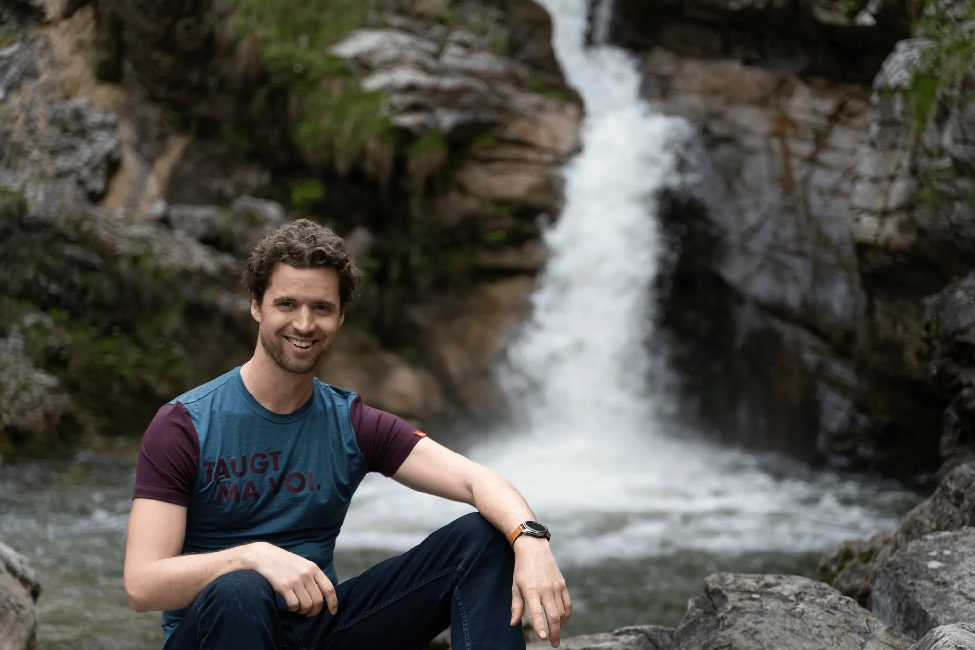 Thilo im Shirt mit Aufdruck „Taugt ma voi.“ vor einem Wasserfall