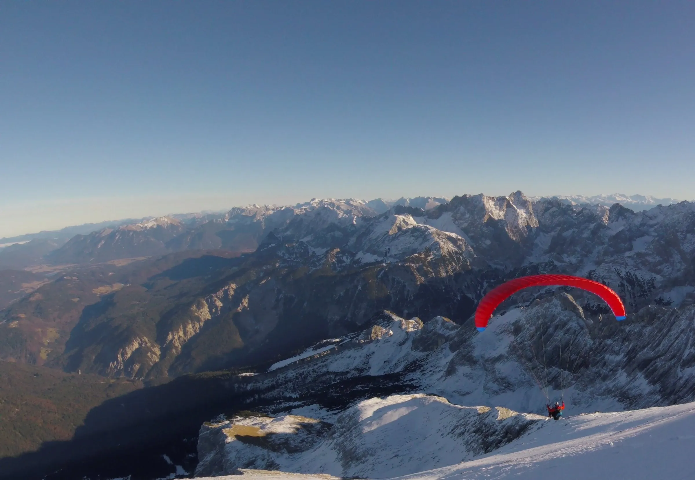Thilo startet mit rotem Gleitschirm an der verschneiten Alpspitze über dem Wettersteingebirge