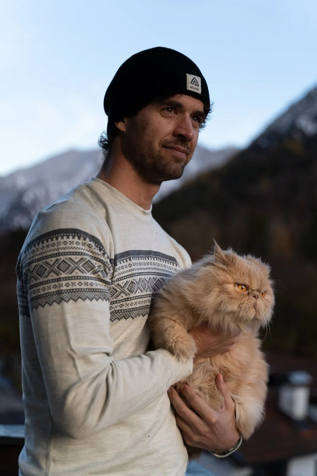 Thilo im Aclima-Merinopullover mit Mütze hält seinen Perserkater vor Bergkulisse