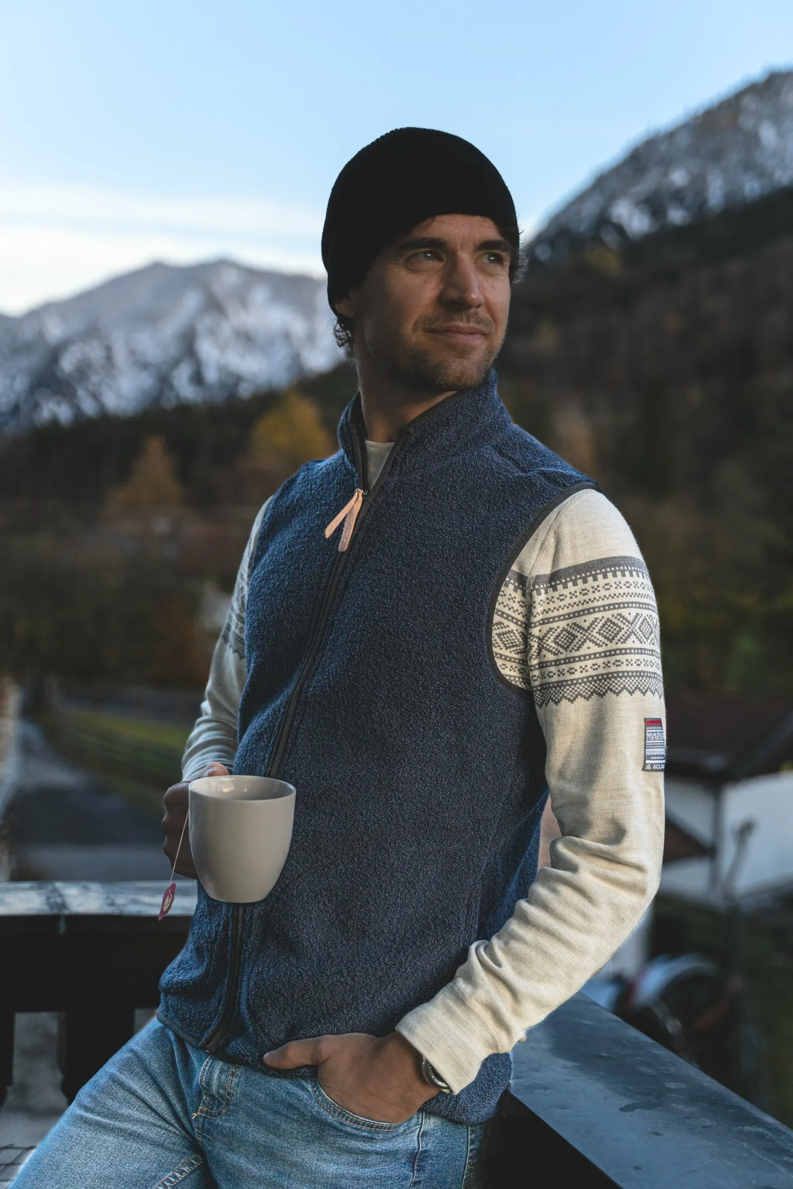 Thilo in Weste und Strickpullover mit Tasse vor herbstlicher Bergkulisse