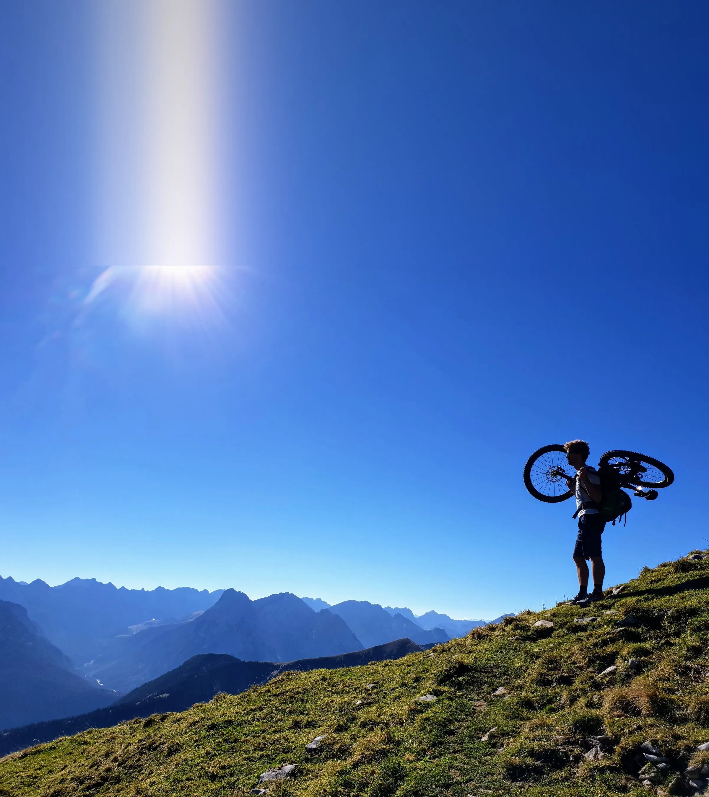 Thilo trägt sein Mountainbike über einen Grasgrat vor blauen Bergketten
