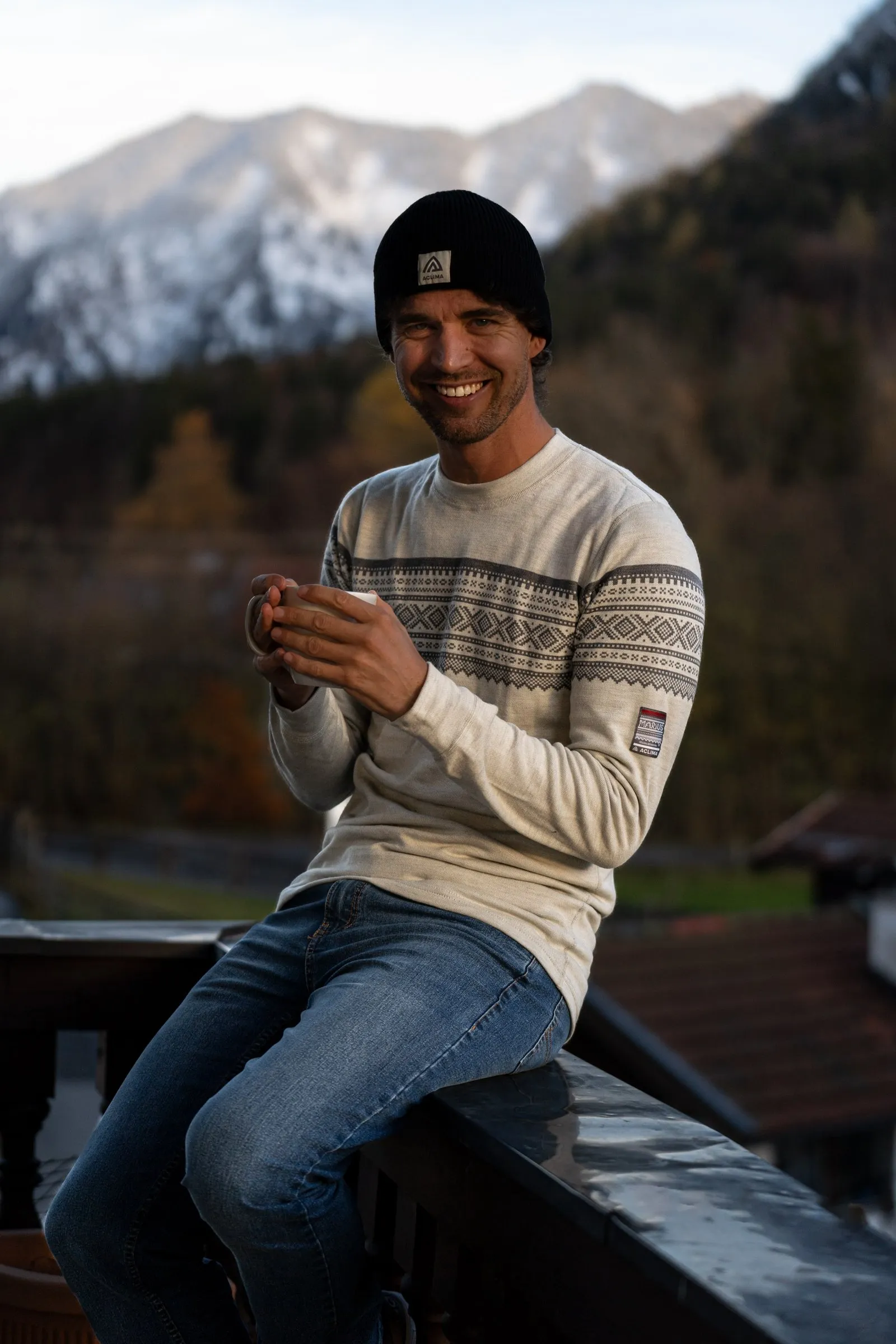 Thilo lacht in die Kamera, mit Mütze, Strickpullover und Tasse vor Bergkulisse